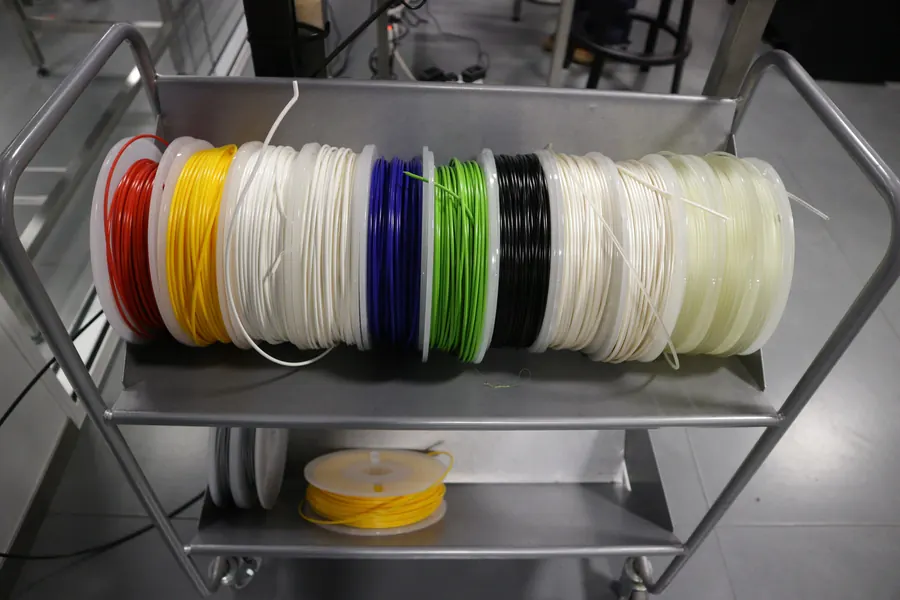 A row of filament spools in red, yellow, white, blue and green lying on an open workshop shelf