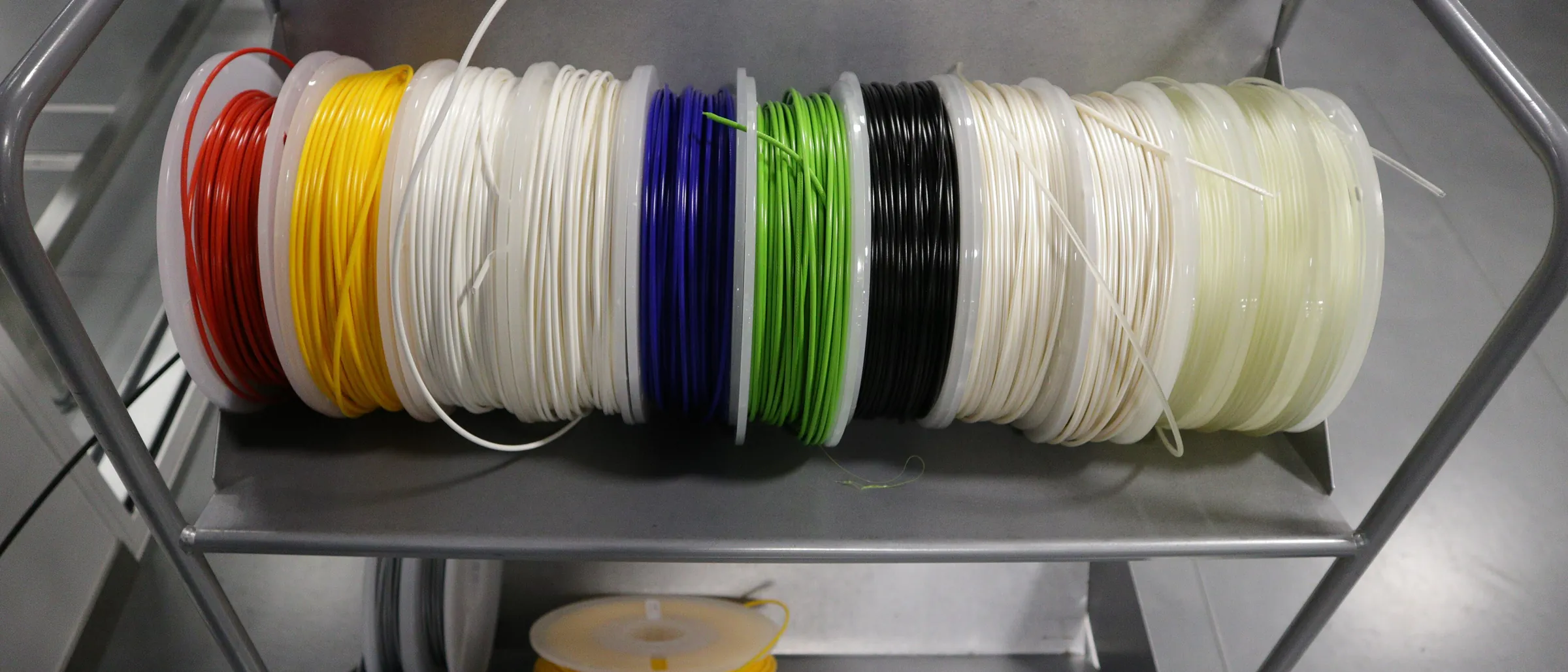 A row of filament spools in red, yellow, white, blue and green lying on an open workshop shelf