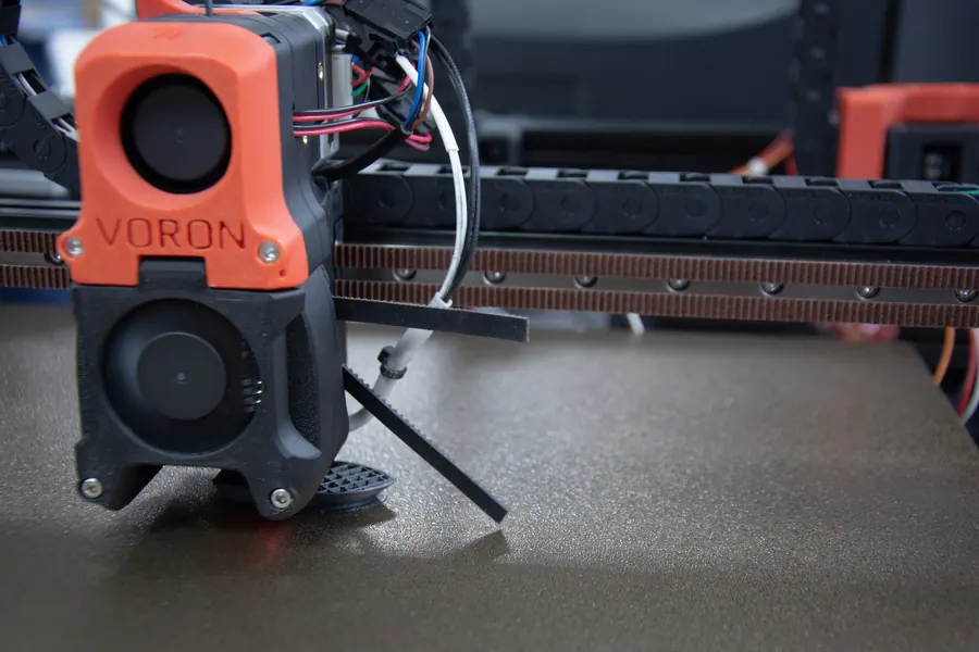 The toolhead of a Voron printer, its orange and black printed housing marked VORON, over a textured build sheet