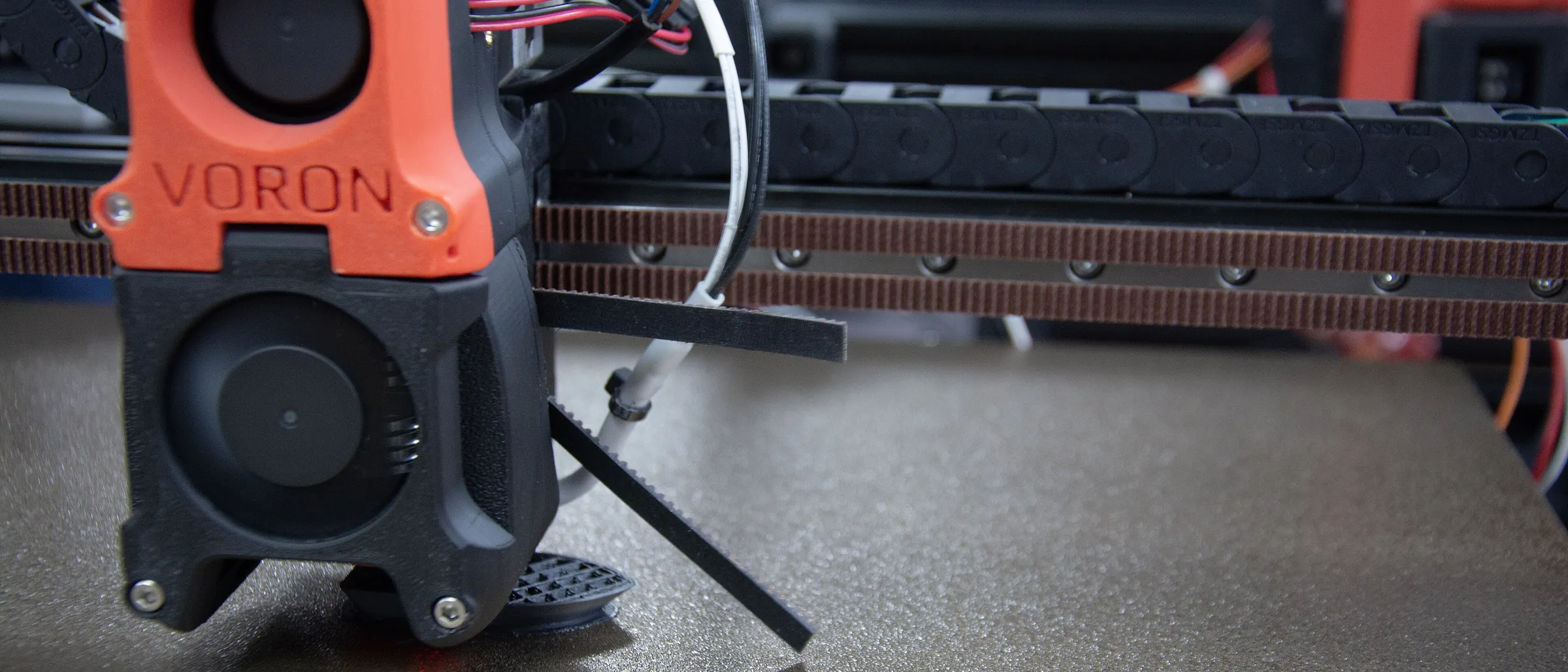 The toolhead of a Voron printer, its orange and black printed housing marked VORON, over a textured build sheet
